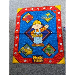 May include: A rectangular blanket featuring Bob the Builder in the center, surrounded by construction vehicles like a dump truck, excavator, and steamroller. The border is red with orange wrench patterns and white screw heads, with the text Bob the Builder.