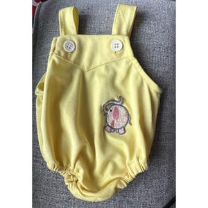 May include: A pale yellow baby romper with white buttons and shoulder straps. It features an embroidered elephant design. The romper has a rounded bottom and is made of a soft fabric.