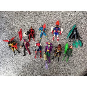 Op de afbeelding: Een verzameling van elf plastic actiefiguren van superhelden en schurken. De set bevat diverse personages met gegoten details, beschilderde kenmerken en bijbehorende plastic accessoires zoals wapens, capes en vleugels in rood, groen en blauw.