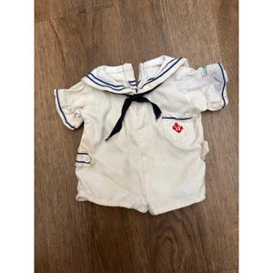 May include: A white short-sleeved sailor-style shirt featuring a large navy blue collar with white trim, a black neck tie, and a small chest pocket adorned with a red embroidered anchor emblem. The garment includes navy blue sleeve cuffs.