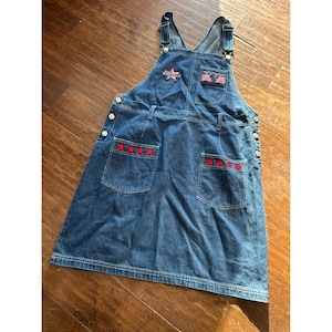Op de afbeelding: Blauwe denim overall jurk met rode en witte ster- en vlagaccenten. Voorzien van verstelbare bandjes, voor- en achterzakken en knoopsluitingen. De zakken zijn versierd met geborduurde rode sterren.