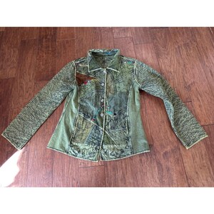 May include: Olive green patchwork jacket with long sleeves. The jacket has a collared neckline, button-down front, and decorative stitching. The fabric has a textured appearance with various patterns and colours.