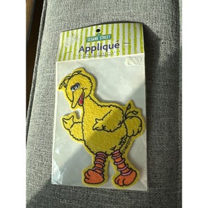 Può includere: Una toppa applicata di Big Bird gialla di Sesame Street. La toppa è in una confezione di plastica trasparente con le parole "Sesame Street Appliqué" stampate in alto. Il personaggio di Big Bird è giallo con gambe e piedi arancioni e un becco rosso aperto.