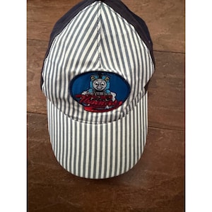 May include: A baseball cap featuring a blue and white vertical pinstripe pattern with solid dark blue side panels. An oval patch on the front displays a blue train character with the red embroidered text Thomas positioned below the train graphic.