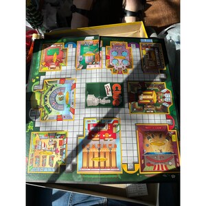 Op de afbeelding: Een Cluedo bordspel met een groen spelbord met verschillende kamers en een centraal Cluedo logo. De kamers zijn geïllustreerd met kleurrijke details, waaronder meubels en objecten. Het spelbord zit in een gele doos.