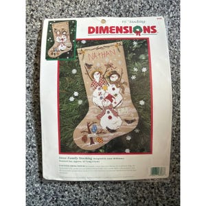 May include: A Dimensions counted cross stitch kit for a 16 inch stocking titled Snow Family Stocking. The packaging features a tan stocking with three snowmen, the name Nathan, and winter motifs including birds, trees, and snowflakes.