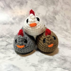 May include: Three crocheted chicken toys in a stack. The top chicken is white, the bottom left is gray, and the bottom right is brown. Each has a red comb, black eyes, and an orange beak. The toys are handmade.