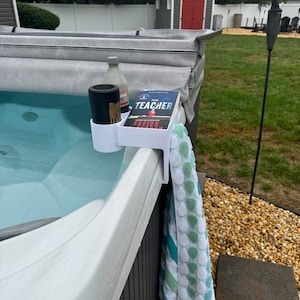 May include: A white plastic hot tub caddy holds a black beverage holder, a bottle, and a book titled "The Teacher" by Freida McFadden. A towel with green and white polka dots hangs on the side of the tub.