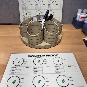 May include: A tan-colored Bourbon Buddy organizer with six cylindrical compartments, each with black horizontal stripes. The organizer sits on a light brown, scalloped base. Two Bourbon Buddy score sheets are visible, with the text "BOURBON BUDDY". Black and white markers are in the organizer.
