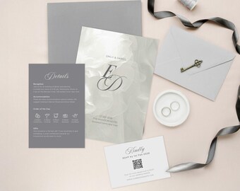 May include: A flat lay of wedding stationery in shades of gray and white. Includes an invitation with initials, a details card, RSVP card, envelopes, a key charm, and a small dish with two rings. A gray ribbon and thread spool are also present.