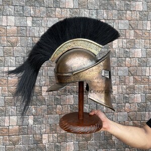 Op de afbeelding: Een bronzen Spartaanse helm met een zwarte paardenhaar pluim, tentoongesteld op een houten standaard. De helm heeft een onderscheidend ontwerp met een verhoogde kam en een gezichtsmasker met een enkele oogopening.