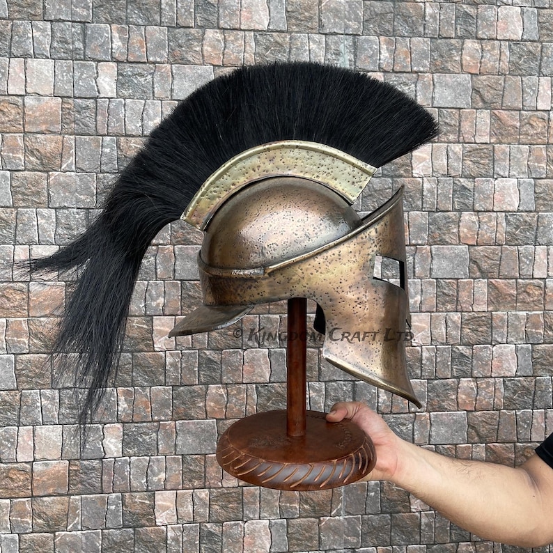 Op de afbeelding: Een bronzen Spartaanse helm met een zwarte paardenhaar pluim, tentoongesteld op een houten standaard. De helm heeft een onderscheidend ontwerp met een verhoogde kam en een gezichtsmasker met een enkele oogopening.