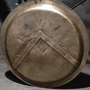 Op de afbeelding: Een rond, bronzen schild met een verhoogd, driehoekig ontwerp in het midden. Het schild heeft een versleten, verweerde uitstraling.