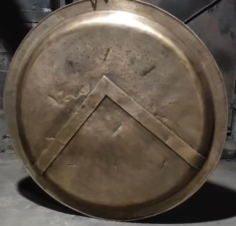 Op de afbeelding: Een rond, bronzen schild met een verhoogd, driehoekig ontwerp in het midden. Het schild heeft een versleten, verweerde uitstraling.