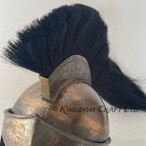 Op de afbeelding: Een bronzen Spartaanse helm met een zwarte paardenhaar pluim. De helm heeft een getextureerd oppervlak en een verhoogde kam. De pluim is lang en vloeiend en is bevestigd aan de helm met een metalen band. De helm is een replica van een historische Spartaanse helm.