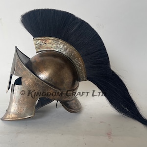 Op de afbeelding: Een bronzen Spartaanse helm met een zwarte paardenhaar pluim. De helm heeft een onderscheidend ontwerp met een verhoogde kam en een gezichtsmasker met een enkele oogopening. De pluim is lang en vloeiend, wat een dramatische toets aan de helm toevoegt.