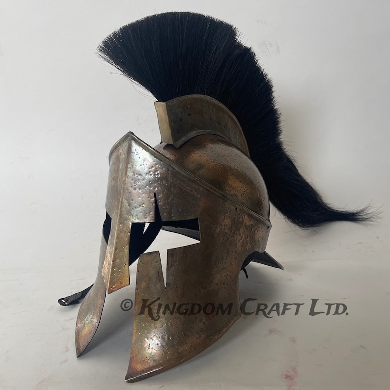 Op de afbeelding: Een bronzen Spartaanse helm met een zwarte paardenhaar pluim. De helm heeft een kenmerkend 'X'-ontwerp aan de voorkant en een getextureerd oppervlak. De helm is gemaakt van metaal en is een replica van een historisch artefact. &copy; KINGDOM CRAFT LTD.