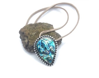 May include: A teardrop-shaped pendant necklace featuring a vibrant abalone shell with iridescent blues and greens, set in a silver-toned bezel. The pendant hangs from a light brown cord, resting on a textured stone against a white background.
