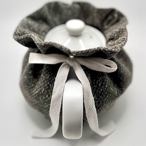 May include: A white ceramic teapot with a lid is nestled inside a gray tweed tea cozy. The cozy is tied with a white ribbon. The teapot handle is visible. The cozy has a ruffled edge.