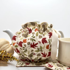 May include: A white ceramic teapot covered by a fabric tea cozy featuring a pattern of red and orange autumn leaves, brown pinecones, and green foliage. The cozy has a gathered top and a cream-colored herringbone fabric handle on the side.