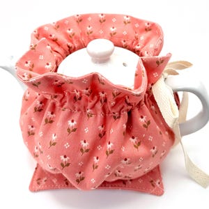 May include: A white ceramic teapot with a matching lid, nestled inside a coral-colored tea cozy. The cozy is adorned with a delicate floral pattern and a cream-colored ribbon tie. A matching coaster sits beneath the teapot.