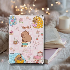 Puede incluir: Una funda transparente para teléfono con un fondo rosa que presenta ilustraciones de carpinchos, flores y los textos "Bookish" y "Just one more chapter". La funda tiene un diseño lindo y caprichoso.
