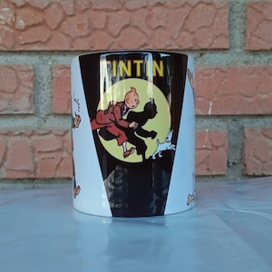 Könnte beinhalten: Zylindrischer Keramikbecher mit einer Cartoon-Illustration von Tim und Struppi. Der Becher hat ein Schwarz-Weiß-Design mit dem Wort "TINTIN" auf der Seite. Der Hintergrund ist eine Backsteinmauer.
