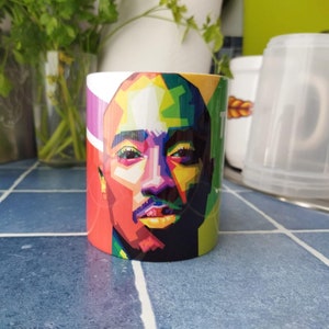Può includere: Una tazza in ceramica bianca con un ritratto astratto e colorato di una persona con la barba nera. Il ritratto è in uno stile che ricorda la pop art.
