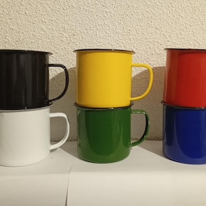 Könnte beinhalten: Sechs Emaille-Becher, paarweise gestapelt. Die oberen Becher sind schwarz, gelb und rot. Die unteren Becher sind weiß, grün und blau.