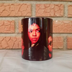 Peut inclure: Une tasse en céramique noire avec un portrait de femme au rouge à lèvres rouge et un collier doré. La tasse affiche également deux autres visages et le mot "S" en or. L'arrière-plan est un mur de briques.