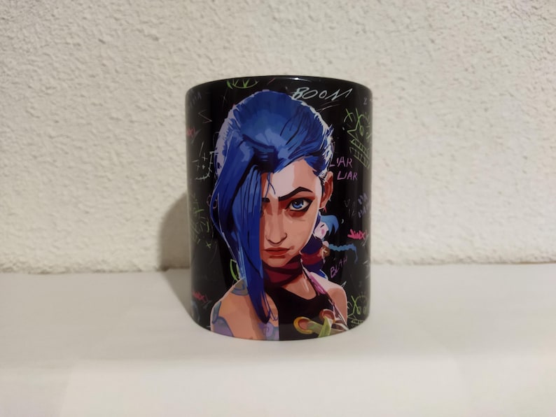 May include: Black ceramic mug with a colorful graphic of a female character with blue hair and a black and pink outfit. The background is black with colorful scribbles and text that includes "BOOM" and "VAR LIAR".