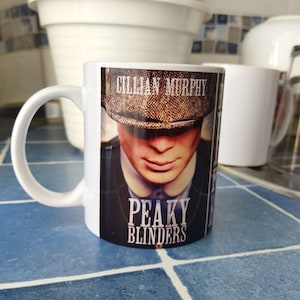 Peut inclure: Une tasse en céramique blanche avec une image en noir et blanc de Cillian Murphy de la série Peaky Blinders. La tasse porte l'inscription "Cillian Murphy" et "Peaky Blinders".