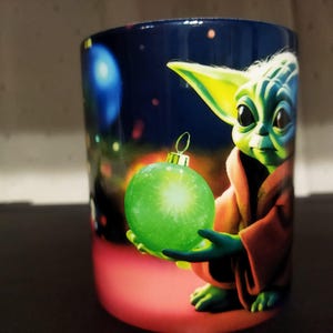 K&ouml;nnte beinhalten: Keramikbecher mit festlichem Design. Er zeigt eine gr&uuml;ne Figur, die eine gr&uuml;ne Kugel h&auml;lt. Der Becher hat einen dunkelblauen Hintergrund mit bunten Lichtern, und die Figur tr&auml;gt eine braune Robe.