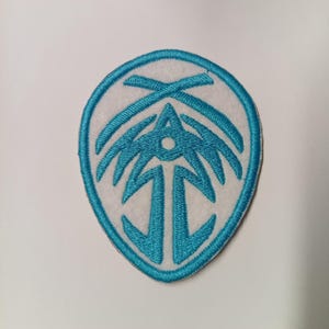 Peut inclure: &Eacute;cusson brod&eacute; avec un fond blanc et un motif bleu sarcelle. Le motif est un embl&egrave;me stylis&eacute; avec un point central et des lignes rayonnantes, enferm&eacute; dans une bordure bleu sarcelle. L'&eacute;cusson est destin&eacute; aux v&ecirc;tements ou accessoires.