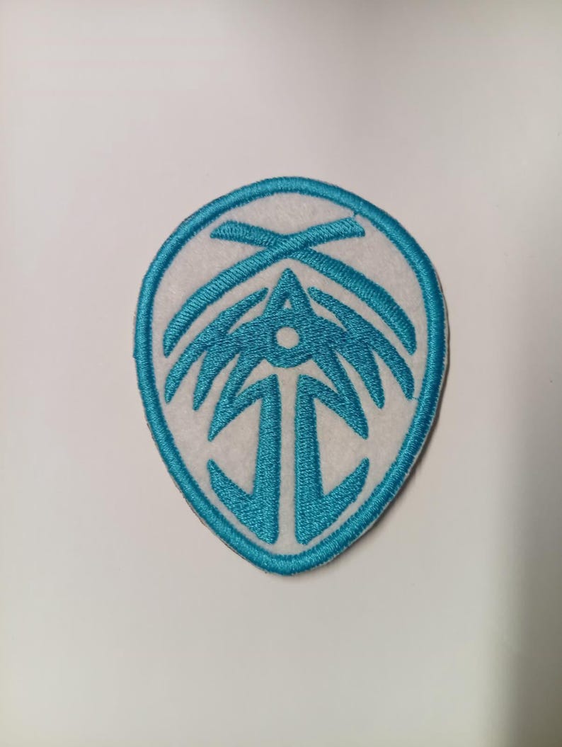 Peut inclure: &Eacute;cusson brod&eacute; avec un fond blanc et un motif bleu sarcelle. Le motif est un embl&egrave;me stylis&eacute; avec un point central et des lignes rayonnantes, enferm&eacute; dans une bordure bleu sarcelle. L'&eacute;cusson est destin&eacute; aux v&ecirc;tements ou accessoires.