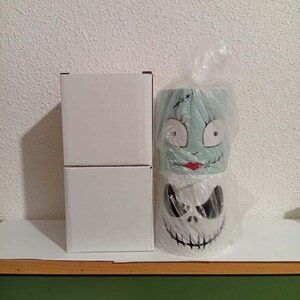 May include: Two ceramic mugs featuring characters from The Nightmare Before Christmas. One mug depicts Sally's face, the other Jack Skellington's. The mugs are in plastic and next to two white boxes.