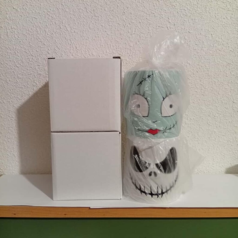 May include: Two ceramic mugs featuring characters from The Nightmare Before Christmas. One mug depicts Sally's face, the other Jack Skellington's. The mugs are in plastic and next to two white boxes.
