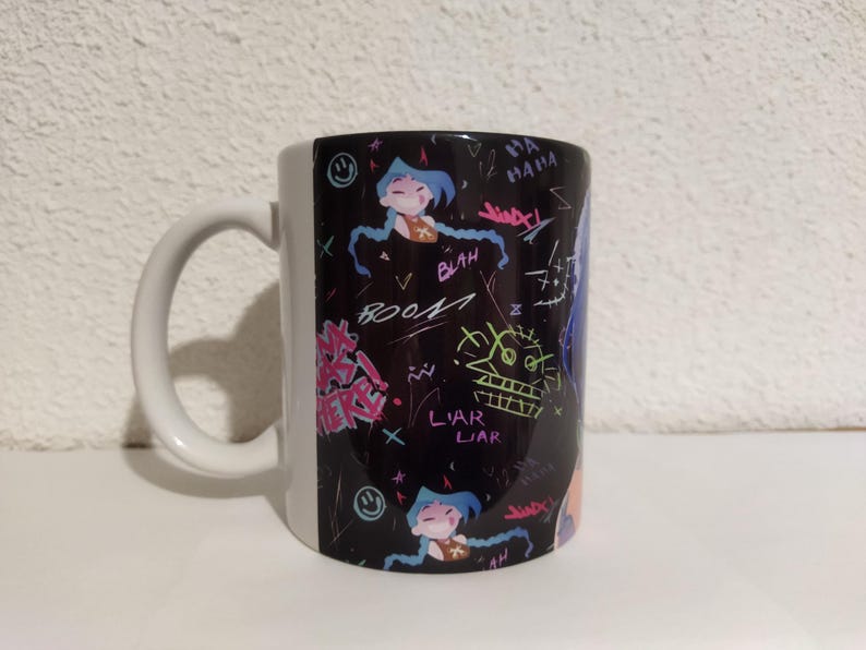 May include: A white ceramic mug with a black handle. The mug has a black and white design with colorful graffiti-style writing and cartoon characters. The writing includes the words "liar liar" and "boo!"