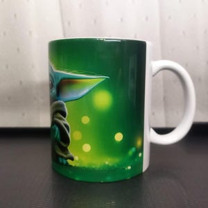 Pu&ograve; includere: Tazza in ceramica bianca con un disegno verde e bianco di un personaggio con grandi orecchie e un mantello verde. La tazza ha un manico bianco e uno sfondo verde con punti gialli luminosi.