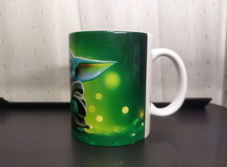 Pu&ograve; includere: Tazza in ceramica bianca con un disegno verde e bianco di un personaggio con grandi orecchie e un mantello verde. La tazza ha un manico bianco e uno sfondo verde con punti gialli luminosi.