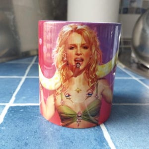 Może przedstawiać: Biały kubek ceramiczny z kolorowym obrazkiem kobiety śpiewającej w mikrofon. Tło to gradient różowego, fioletowego i żółtego. Kobieta ma na sobie zielono-złoty top i blond włosy.