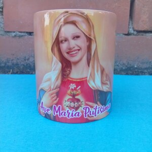May include: A cylindrical ceramic mug with a portrait of a woman with a halo and a red top with a heart design. The mug has the text "Ave Maria Putisima" in cursive. The background is a light peach colour.