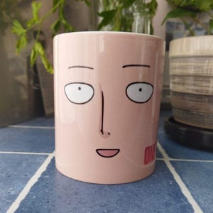 Puede incluir: Una taza de cerámica rosa con una cara de dibujos animados y la palabra "ONE" impresa en la parte inferior. La cara tiene ojos grandes, una nariz y una boca sonriente.