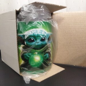 Pu&ograve; includere: Una tazza di ceramica verde con un personaggio dei cartoni animati con grandi occhi e orecchie a punta, che tiene in mano un globo verde brillante. La tazza &egrave; all'interno di una scatola di cartone, parzialmente avvolta nella plastica.