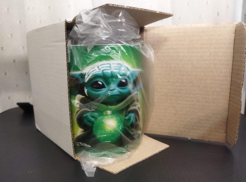 Pu&ograve; includere: Una tazza di ceramica verde con un personaggio dei cartoni animati con grandi occhi e orecchie a punta, che tiene in mano un globo verde brillante. La tazza &egrave; all'interno di una scatola di cartone, parzialmente avvolta nella plastica.