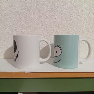 May include: Two ceramic mugs with character faces. One is white with a black face design, and the other is light blue with a black face design. Both mugs have white handles and are on a white surface.