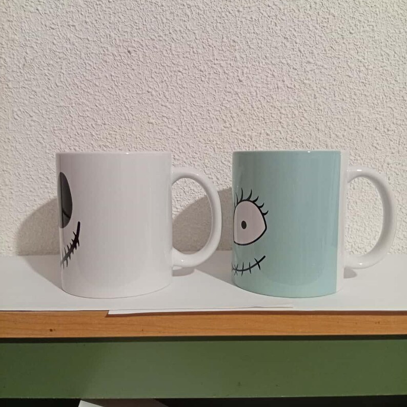 May include: Two ceramic mugs with character faces. One is white with a black face design, and the other is light blue with a black face design. Both mugs have white handles and are on a white surface.