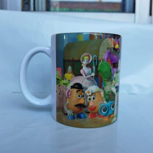 May include: White ceramic mug featuring a colorful design with characters from the animated film Toy Story. The design includes Woody, Buzz Lightyear, and other characters. The mug has a white handle and a glossy finish.