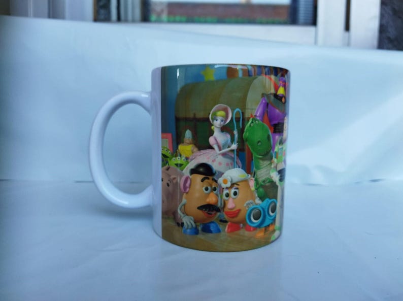 May include: White ceramic mug featuring a colorful design with characters from the animated film Toy Story. The design includes Woody, Buzz Lightyear, and other characters. The mug has a white handle and a glossy finish.