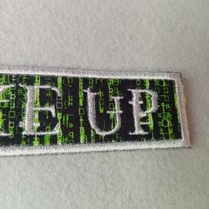 Pode incluir: Patch retangular com a palavra "GEE UP" bordada em fio prateado. O fundo &eacute; preto com um padr&atilde;o de chuva digital verde. O patch tem uma borda branca.